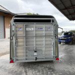 Rear view of a silver livestock trailer in a loading bay, with a blue pickup truck and a person in the background on the right.