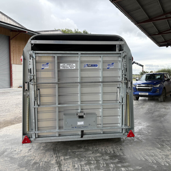 Rear view of a silver livestock trailer in a loading bay, with a blue pickup truck and a person in the background on the right.