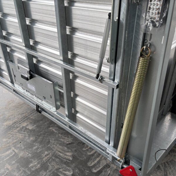 Close-up of a metal trailer gate with a vertical hydraulic strut, coil springs, and a control keypad mounted on the side panel.
