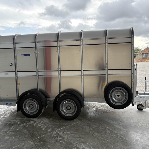 Long silver multi-axle livestock trailer with riveted panels, parked on a concrete lot under a cloudy sky.