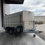 Silver Ifor Williams trailer parked inside a warehouse bay beside a corrugated metal wall and gravel yard beyond.