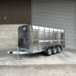 Silver enclosed trailer with a curved front parked against a brown corrugated metal wall and a large gray roll-up door behind it.