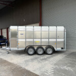 Silver trailer hitched to a blue pickup truck, parked beside a large corrugated metal warehouse with a closed rolling shutter door.
