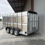 A silver livestock trailer with a curved roof sits on a gravel lot beside a corrugated metal building; a green tractor is visible in the background on the left.