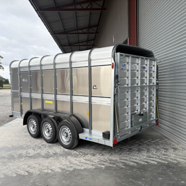 A silver livestock trailer with a curved roof sits on a gravel lot beside a corrugated metal building; a green tractor is visible in the background on the left.