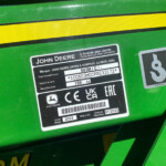 Close-up of a John Deere equipment sticker on a green surface, showing model and serial information next to a yellow reflective stripe.