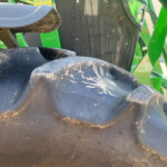Close-up of a dirty black tractor tire with green farming equipment in the background, mud specks on the tread.