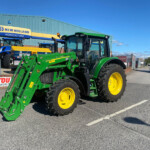 Green tractor with a front loader and large yellow wheels parked outside a New Holland Agriculture building, with blue tractors in the background on a sunny day.