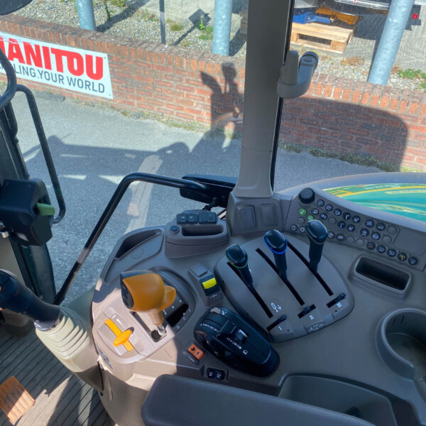 Driver's seat view of a tractor cab with multiple levers, knobs, and a joystick, brick wall and sign visible through the window outside