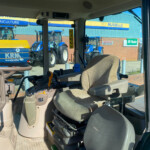 Interior of a tractor cab with beige seat and control levers, looking out at blue tractors parked outside.