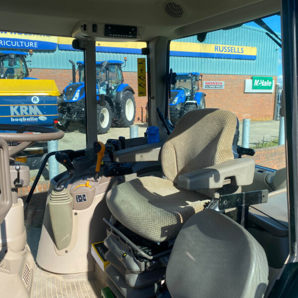 Interior of a tractor cab with beige seat and control levers, looking out at blue tractors parked outside.