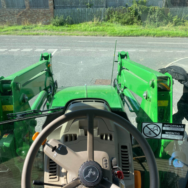 View from inside a green tractor cab, looking at a street with a grassy verge and a brick wall beyond; loader arms frame the sides.