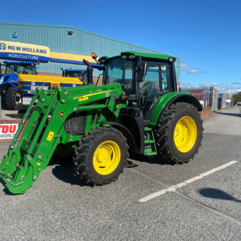 Green tractor with a front loader and large yellow wheels parked outside a New Holland Agriculture building, with blue tractors in the background on a sunny day.