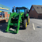 Green John Deere tractor with front loader in a parking lot beside a brick building and blue sky, with Russell's sign in the background.