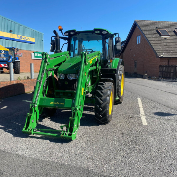 Green John Deere tractor with front loader in a parking lot beside a brick building and blue sky, with Russell's sign in the background.