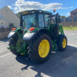 Green tractor with large yellow wheels parked on a residential street, blue sky with clouds overhead and houses with fences in the background
