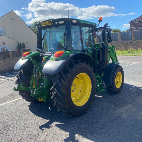 Green tractor with large yellow wheels parked on a residential street, blue sky with clouds overhead and houses with fences in the background