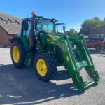 Green John Deere tractor with front loader parked on a residential street, blue sky and brick houses in the background, yellow wheels visible in the foreground as a loader attachment hangs off the front.