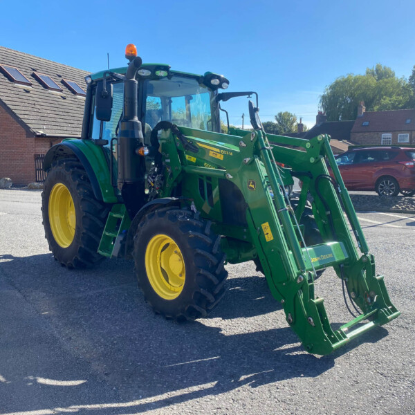 Green John Deere tractor with front loader parked on a residential street, blue sky and brick houses in the background, yellow wheels visible in the foreground as a loader attachment hangs off the front.