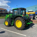 Green tractor with a front loader and yellow wheels parked in a lot near an industrial building under a blue sky, right side.