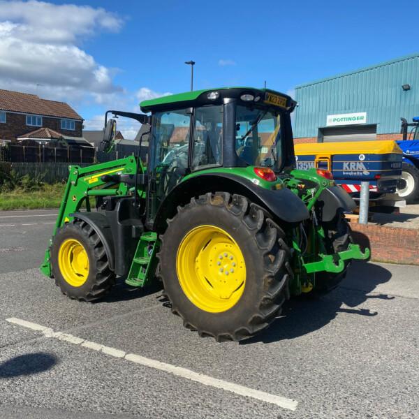Green tractor with a front loader and yellow wheels parked in a lot near an industrial building under a blue sky, right side.