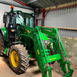 Green John Deere tractor with a front loader attachment parked inside a metal shed, orange beacon on the cab roof, large rear tire visible.