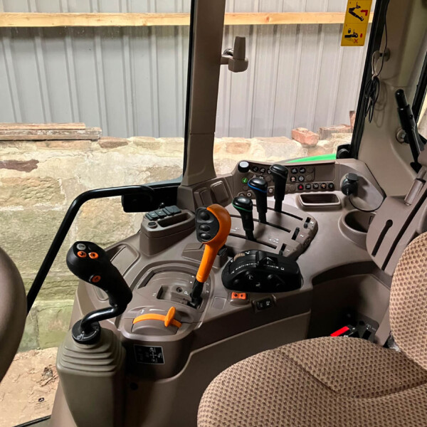 Interior view of a construction vehicle cabin with multiple joysticks and levers on a gray console, beige seat visible, outside a brick wall and metal siding.