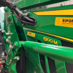 Close-up of a green agricultural machine showing a hydraulic hose assembly attached to a mounting bar, with safety labels nearby.