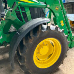Close-up of a green John Deere tractor with a large yellow wheel, hydraulic arm, and black tire tread in a workshop setting.