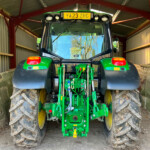 Rear view of a green John Deere tractor with oversized muddy tires inside a metal shed; yellow license plate YA23 ZRG on the back.