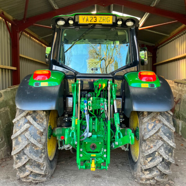 Rear view of a green John Deere tractor with oversized muddy tires inside a metal shed; yellow license plate YA23 ZRG on the back.
