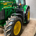 Green John Deere tractor with large yellow wheels, parked indoors beside a metal roll-up door, showing front loader attachment and cab window reflections.