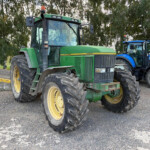 Two large tractors parked side by side in a lot with trees in the background; green John Deere and blue New Holland models visible.