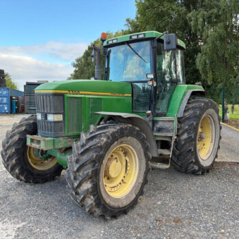 Front 3/4 view of a green John Deere tractor with large muddy rear tires in a gravel yard, blue containers in the background.