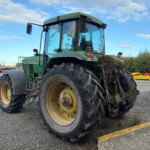 Green agricultural tractor with oversized rear tires parked in a gravel lot; red dumpsters and other farm equipment visible in the background on a sunny day.