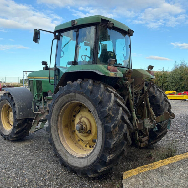 Green agricultural tractor with oversized rear tires parked in a gravel lot; red dumpsters and other farm equipment visible in the background on a sunny day.