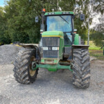 Front view of a green tractor with oversized tires parked on gravel, with trees and fencing in the background