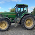 Close-up side view of a green John Deere 7700 tractor with large yellow wheels parked on a gravel lot under a blue sky.