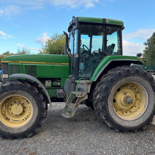 Close-up side view of a green John Deere 7700 tractor with large yellow wheels parked on a gravel lot under a blue sky.