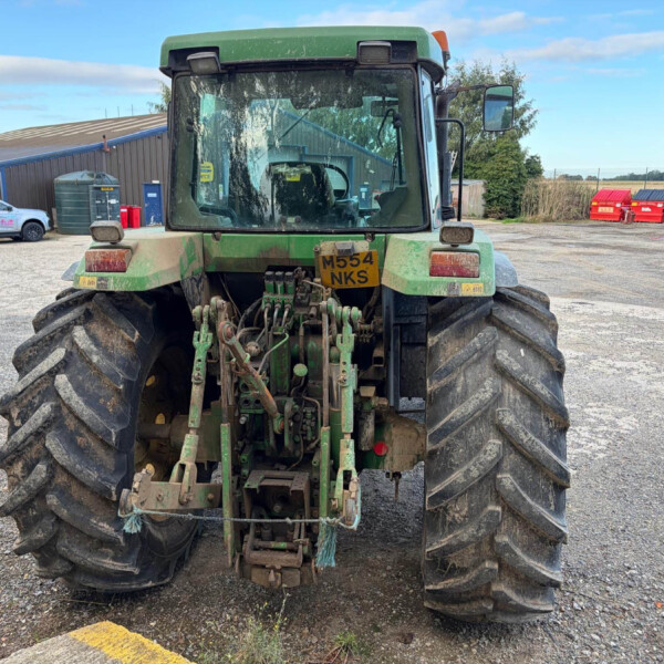 Rear view of a green tractor with large, muddy tires and hooked implements, parked on a gravel lot facing away from the camera.