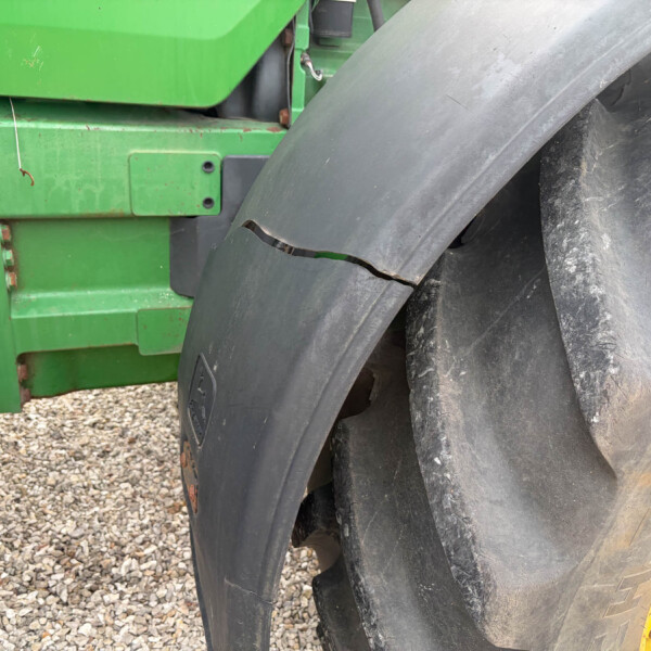 Close-up of a green agricultural machine frame beside a large, weathered tractor tire on a gravel surface, showing cracks in the tire tread.