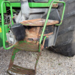 Close-up of rusted metal steps attached to a green tractor, next to a large black tire on gravel ground with yellow wheel rim nearby
