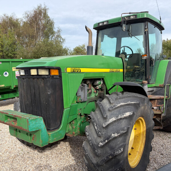 John Deere 8100 tractor with large rear tires and a green body, parked on a gravel lot with a green trailer in the background.