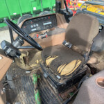 Interior of a worn farm tractor cab with a torn brown seat, steering wheel, and a detailed control panel; outside, large green machinery is visible through the window.