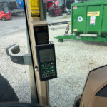 Interior view of a tractor/cab with a control panel and screen, looking out at a gravel yard with a green trailer outside