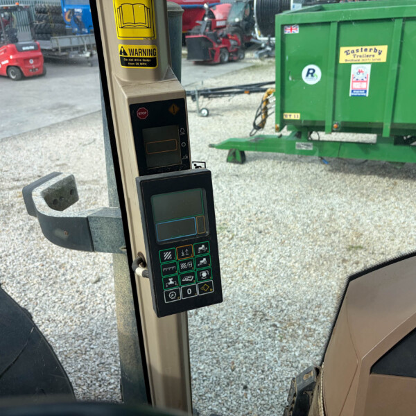 Interior view of a tractor/cab with a control panel and screen, looking out at a gravel yard with a green trailer outside