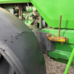 Close-up of a large black tractor tire with a green farming machine in the background and rusted metal brackets nearby on a gravel surface.