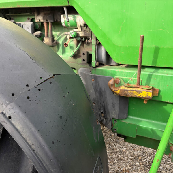 Close-up of a large black tractor tire with a green farming machine in the background and rusted metal brackets nearby on a gravel surface.