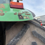 Close-up of a green tractor rear with peeling paint, red taillight, and a muddy, oversized wheel in an industrial yard.