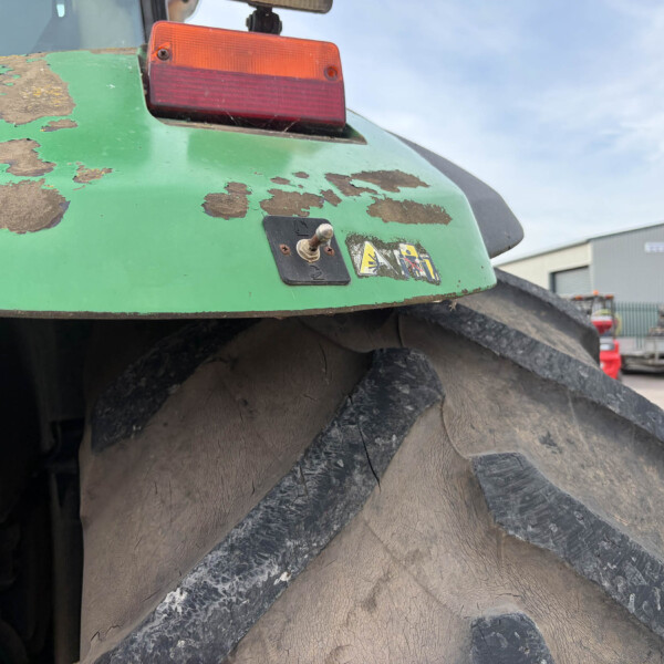 Close-up of a green tractor rear with peeling paint, red taillight, and a muddy, oversized wheel in an industrial yard.
